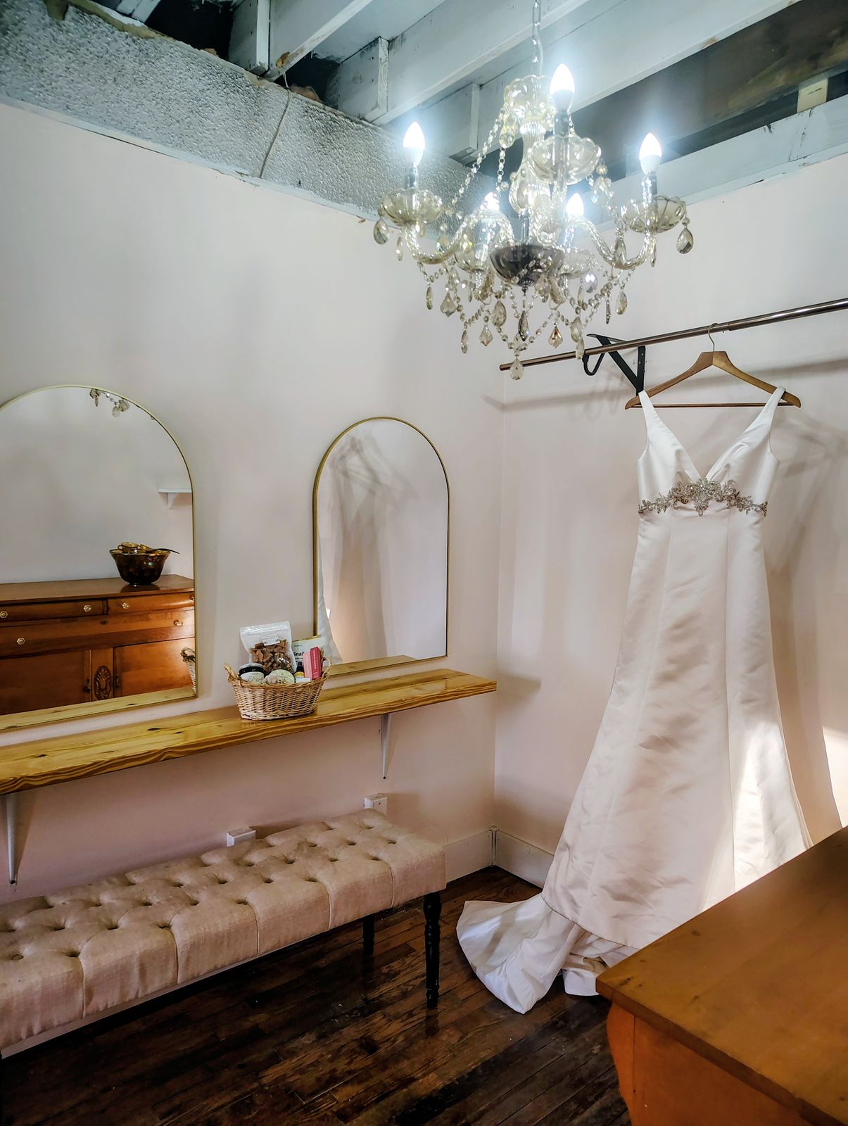 The bridal suite with a wedding gown hanging beside an arched mirror, a tufted bench and a wooden dresser