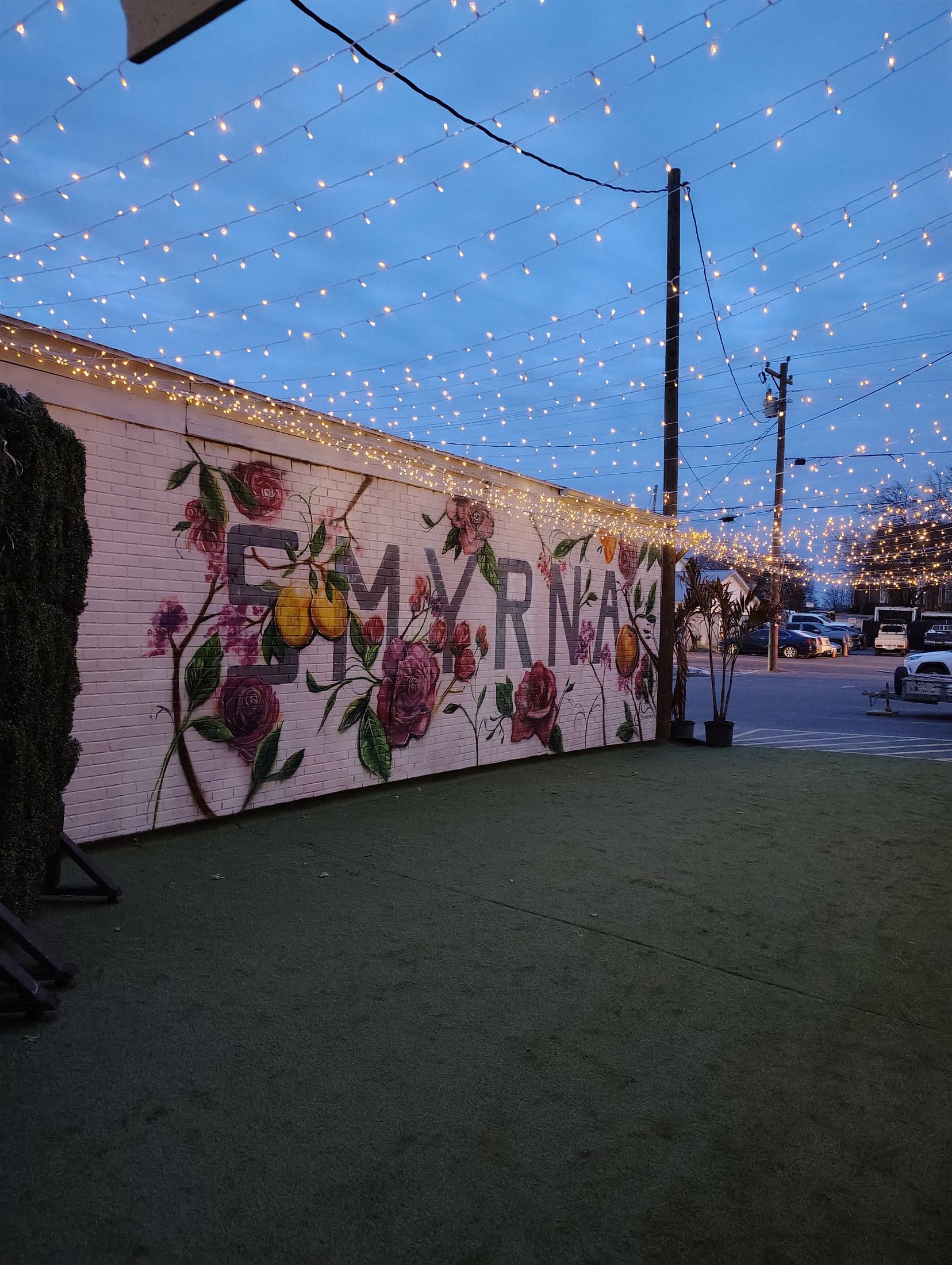 The courtyard at Downtown Social at dusk, strung overhead with warm bulbs above a white brick wall painted with roses and the word Smyrna