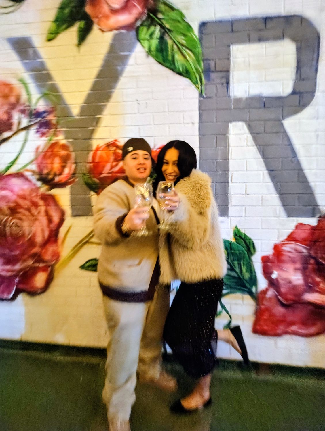 Two guests standing together on the courtyard turf in front of the painted rose mural, holding drinks