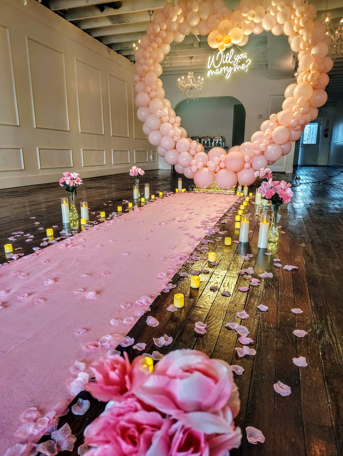 A blush aisle runner scattered with rose petals leading to a balloon heart arch and a neon sign reading will you marry me