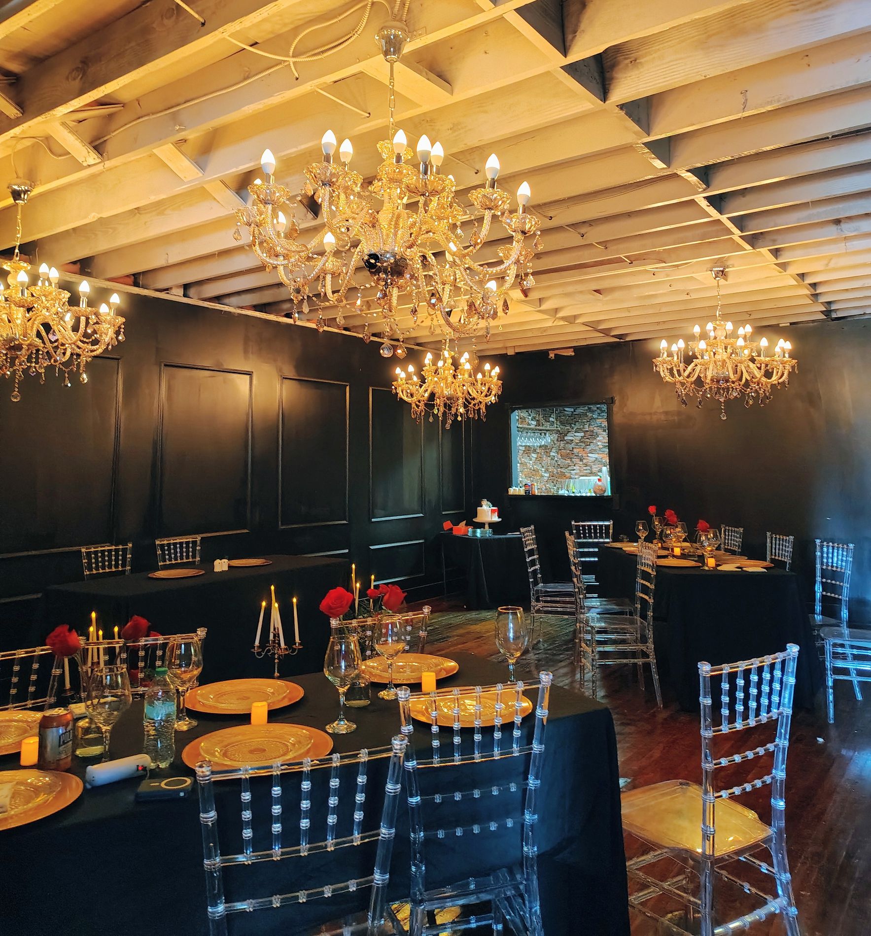 The Speakeasy set for dinner with black linens, gold charger plates, red roses and candlesticks, under three brass chandeliers against glossy black paneled walls