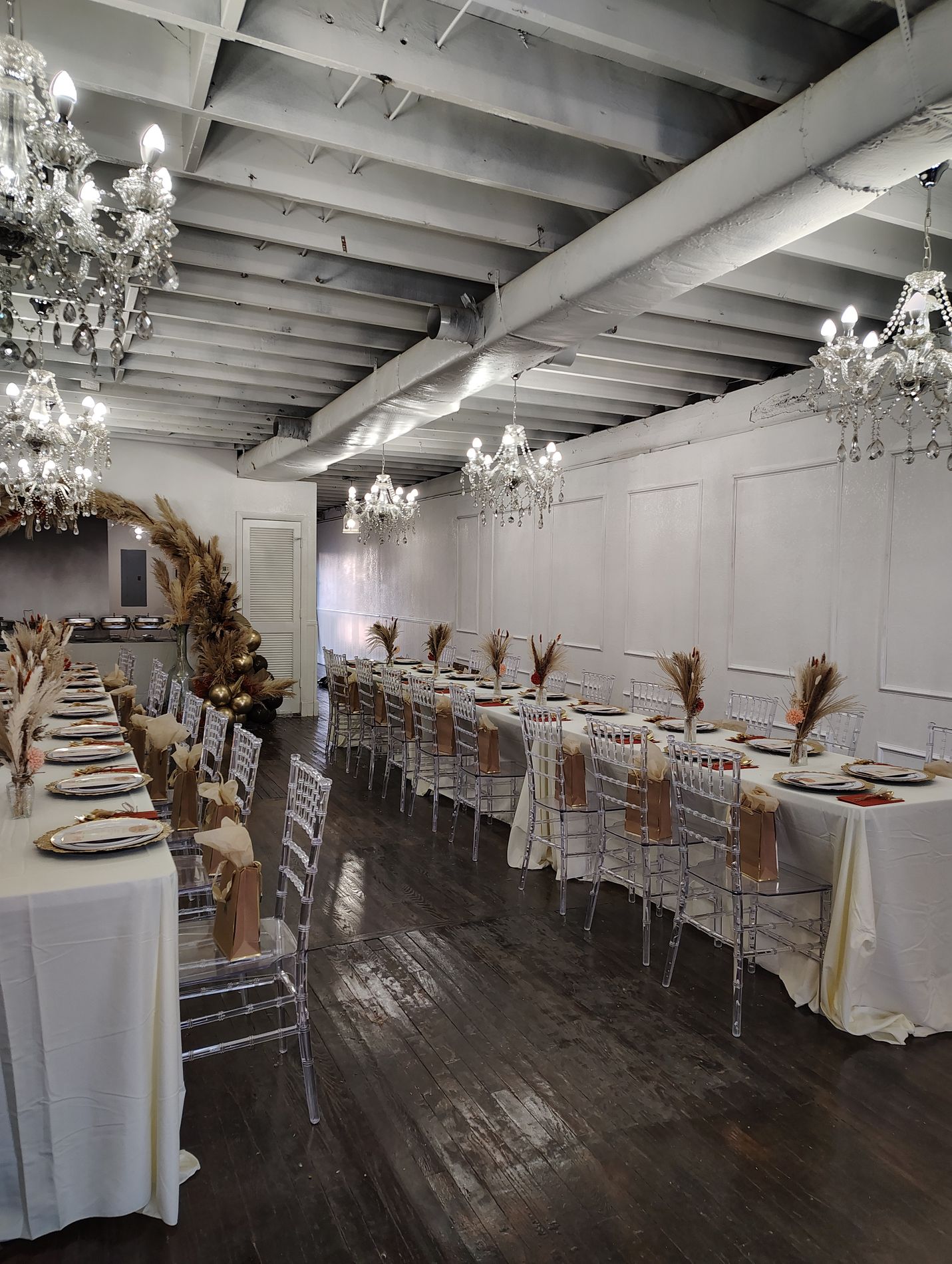 The Traditional Room set with long tables in white linen, clear chiavari chairs and dried floral arrangements, under six crystal chandeliers hung from white painted beams